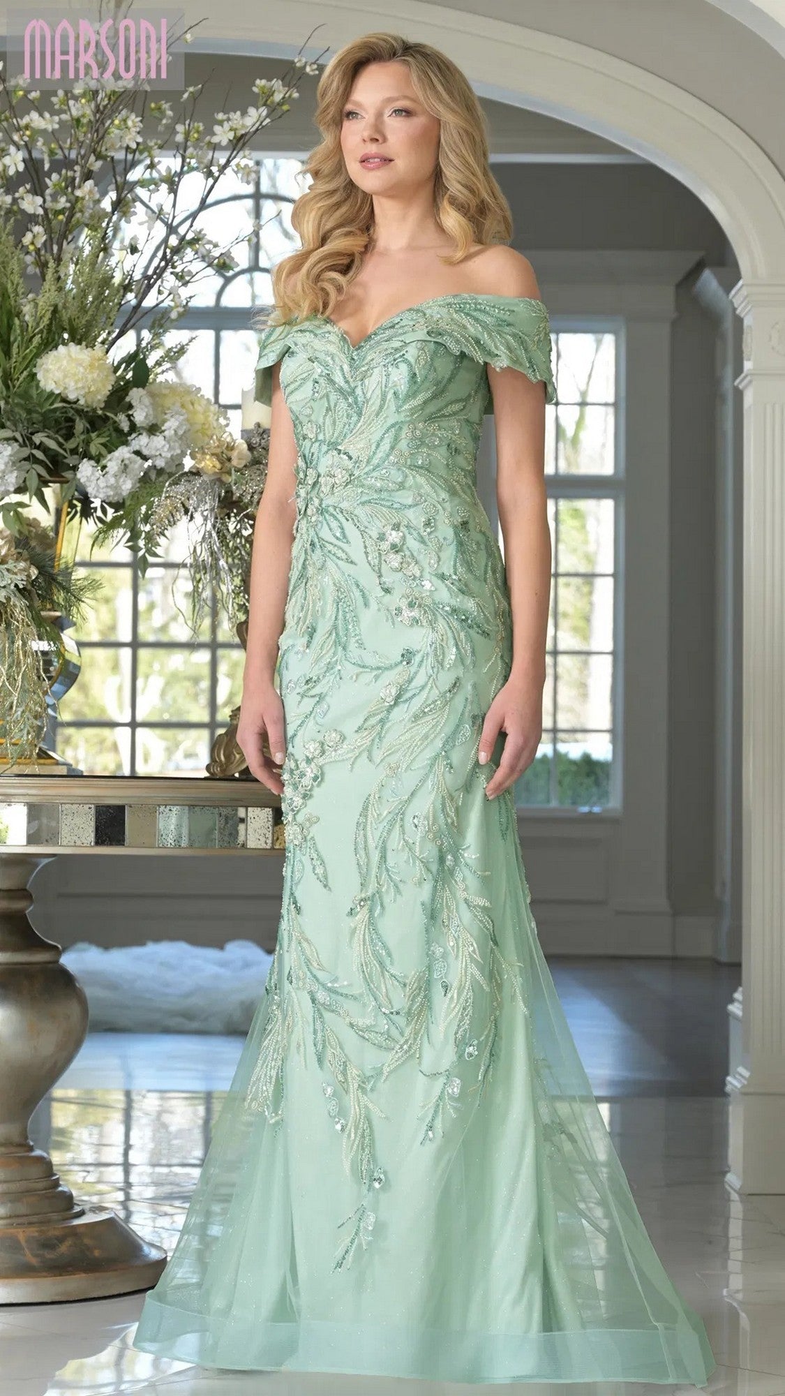 A woman stands indoors in the Marsoni MV1372 long formal dress in spearmint, featuring an off-shoulder design, beaded embroidery, and a fitted bodice that flares gracefully beside a table with a large floral arrangement.